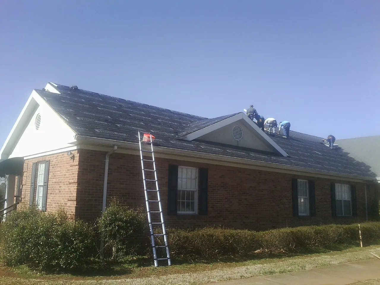 Crew tearing shingles off a brick ranch roof with a ladder set — Metal Roof Repair work by our crew in Lena